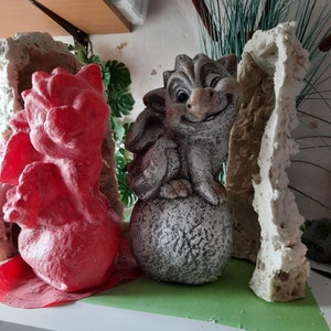 May include: A grey stone dragon figurine with a winking face, sitting on a round stone base. The dragon is surrounded by two red and white moulds.