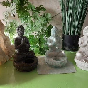 Puede incluir: Tres pequeñas estatuas de Buda, una negra, una blanca y una azul claro, están dispuestas sobre una superficie verde. Cada estatua está sentada en posición de loto con las manos juntas en oración. Las estatuas están sobre pequeñas bases con un espacio hueco para una vela o incienso.