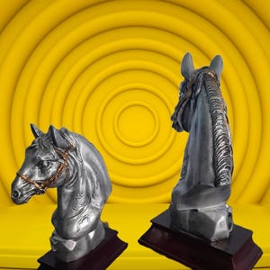 Puede incluir: Dos esculturas de cabeza de caballo de plata sobre bases de madera. Los caballos están mirando en direcciones diferentes. Las esculturas están sobre un fondo amarillo.