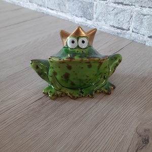 Könnte beinhalten: Eine grüne Keramik-Froschfigur mit braunen Flecken, die eine goldene Krone trägt. Der Frosch hat große, weiße Augen und ein breites, zahniges Grinsen.