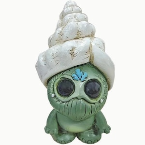May include: A green ceramic figurine with a white seashell hat. The figurine has large eyes, a beard, and a blue flower on its forehead.