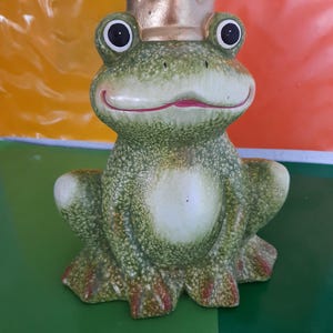 May include: A ceramic frog ornament wearing a gold crown. The frog is predominantly green with white and red detailing. It features large black eyes and a smiling red mouth. The ornament is designed to sit upright.
