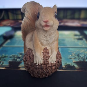 May include: A detailed figurine of a squirrel, featuring a light brown body and a fluffy tail. The squirrel is perched on a textured, brown base, possibly a pine cone. The figurine has a smiling expression and is a decorative item.