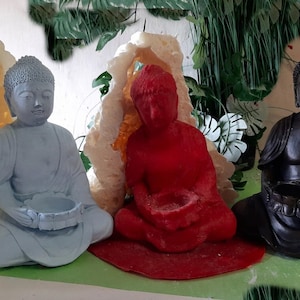 Può includere: Tre statue di Buddha, una bianca, una rossa e una nera, ciascuna con una ciotola in grembo. Le statue sono sedute su una superficie verde con uno sfondo bianco.