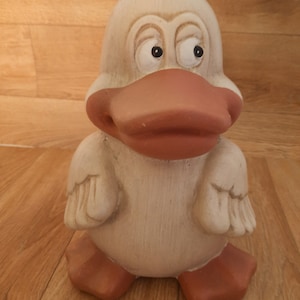 Könnte beinhalten: Eine Keramik-Entenfigur mit weißem Körper, braunem Schnabel und orangefarbenen Füßen. Die Ente hat große, runde Augen und einen skurrilen Ausdruck.