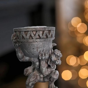 May include: A gray stone gargoyle sculpture holding a round planter with a decorative rim. The planter is empty and the gargoyle has a fierce expression.