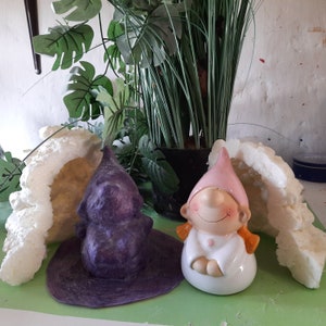 May include: A pair of garden gnome ornaments. One is a purple gnome candle, the other a ceramic gnome with a pink hat and smiling face. Artificial plants and foam decorations are in the background. A charming garden decor item.