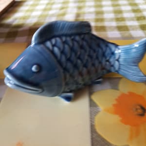 Puede incluir: Una figurita de pescado de cerámica azul con escamas detalladas y una aleta. El pez tiene un acabado esmaltado y una apariencia realista.