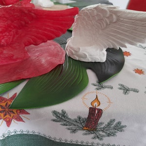 May include: Two decorative angel wings, one red and one white, are displayed on a green leaf-shaped surface. The wings are made of a smooth, glossy material and have a realistic appearance. The wings are placed on a white tablecloth with a green and red star pattern.