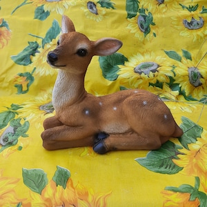 Puede incluir: Una figurita de cervatillo marrón y blanco con manchas, tumbada de lado con la cabeza girada hacia la derecha. La figurita está sobre un fondo floral amarillo y verde.