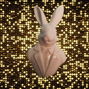 Puede incluir: Busto de conejo blanco de cerámica con traje. El conejo tiene orejas largas y una expresión sonriente. El busto está sobre un fondo brillante dorado.
