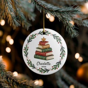 May include: A round white ceramic ornament hanging by a gold string. The centre features a stack of books shaped like a Christmas tree topped with a gold star. A wreath of green pine branches and red berries surrounds the books. The text reads Charlotte 2026.