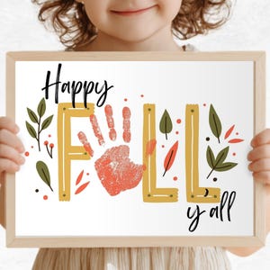 Puede incluir: Un cartel enmarcado con las palabras "Happy Fall y'all" en escritura negra. La palabra "FALL" está en letras amarillas con una huella de mano en el centro. Ilustraciones de hojas y bayas verdes y rojas rodean el texto.