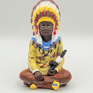 Könnte beinhalten: Eine kleine Figur eines Indianers, der ein gelbes Hemd, eine braune Hose und ein buntes Kopfschmuck trägt. Er hält eine Pfeife in seiner rechten Hand und sitzt mit gekreuzten Beinen.