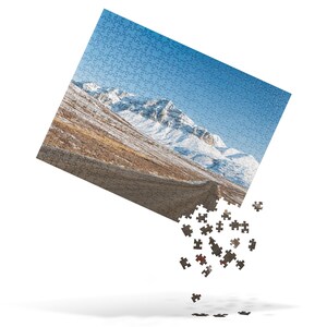 May include: A jigsaw puzzle with a scenic image of a snow-covered mountain range and a winding road. The puzzle is partially assembled, with some pieces scattered around the image.