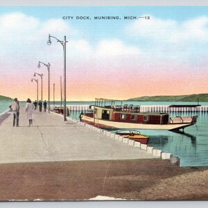 May include: A vintage postcard showing the City Dock in Munising, Michigan. The scene features a long concrete pier with tall street lamps, a boat docked alongside, and people walking on the pier under a soft, pastel-colored sky at sunset.
