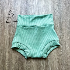 May include: Mint green baby shorts with a ribbed texture. The high-waisted shorts are made of a soft fabric and are designed for comfort. The shorts are displayed on a wooden surface. The logo "Wandering Things Co." is in the upper left corner.