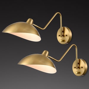 May include: Two brass-colored wall sconces with a curved arm and a dome-shaped shade. The shades have a white interior, and the sconces are mounted on round backplates. The design is a mid-century modern style.