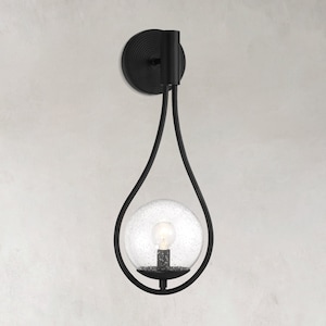 May include: Black metal wall sconce with a clear glass globe shade. The sconce has a teardrop shape and is attached to the wall with a round backplate.