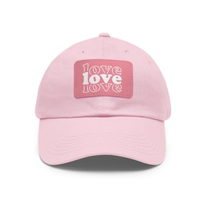 May include: A light pink baseball cap with a curved brim. The cap features a rectangular patch with the word "love" stacked three times in white, set against a pink background. The hat is a casual accessory.