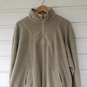 May include: A beige fleece pullover with a quarter zip closure, a drawstring hem, and two side pockets.