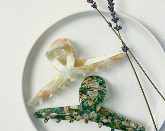 May include: Two hair claw clips. One is a light green and cream colour, and the other is green with a speckled pattern. The clips are on a white plate with sprigs of lavender.