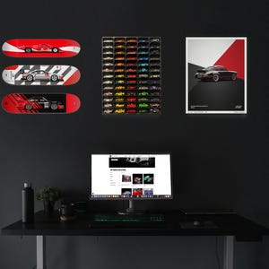 May include: A black computer desk with a black computer monitor, a black keyboard, a black mouse, a black water bottle, a green plant, and a black mug. Three skateboards with car graphics are hanging on the wall behind the desk. A framed poster with a black car on a red and black background is also hanging on the wall. A wall display of miniature cars is on the wall to the left of the computer monitor.