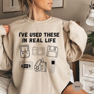 May include: A beige sweatshirt with the text "I've used these in real life" printed on the front. The text is surrounded by images of a handheld video game, a camera, a floppy disk, a VHS tape, headphones, and a cell phone.