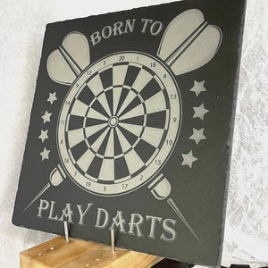 Könnte beinhalten: Ein schwarzes Schiefer-Schild mit dem Text "BORN TO PLAY DARTS" in weißen Buchstaben. Das Schild zeigt eine Dartscheibe mit einem weißen Bullseye und schwarzen und weißen Segmenten. Zwei Darts sind hinter der Dartscheibe gekreuzt.
