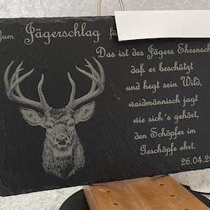 Puede incluir: Placa de pizarra negra con texto grabado y cabeza de ciervo. El texto incluye frases en alemán y la fecha 26.04.2025. La placa es rectangular y tiene un colgador de metal.