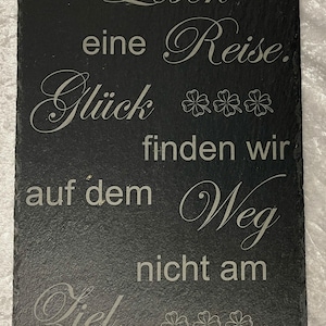 Può includere: Un cartello rettangolare in ardesia nera con testo bianco inciso. Il testo recita "Das Leben ist eine Reise. Glück finden wir auf dem Weg nicht am Ziel." con disegni di trifogli. Un oggetto decorativo.