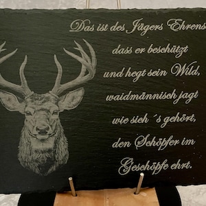 Könnte beinhalten: Schwarze Schiefertafel mit detaillierter Gravur eines Hirschkopfes und Geweihs. Die Tafel zeigt eine elegante Schrift in Silber mit deutschem Text. Der Text übersetzt sich in ein Jägerbekenntnis. Die Tafel wird auf einem Ständer präsentiert.