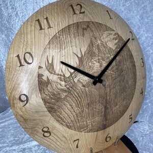 Puede incluir: Un reloj de madera con una escena de ciervo y montaña grabada en la cara. El reloj tiene manecillas negras y marcas de números romanos.