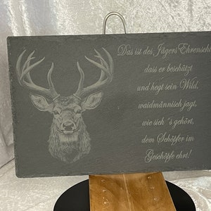 Puede incluir: Una placa de pizarra negra con una cabeza de ciervo grabada en ella. La placa tiene un colgador plateado y el texto "Das ist des Jägers Ehrenschild, dass er beschützt und hegt sein Wild, waidmännisch jagt, wie sich's gehört, dem Schöpfer im Geschöpfe ehrt!" está grabado en ella.