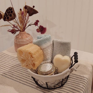 Op de afbeelding: Een zwarte draadmand gevuld met bad- en lichaamsverzorgingsproducten. De mand bevat een loofah, twee handdoeken, een hartvormige zeep en een pot badzout. De mand staat op een gestreept wit en grijs tafelkleed.