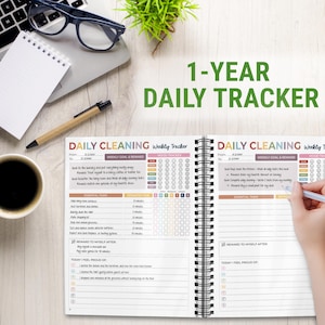 Pode incluir: Um planejador de limpeza di&aacute;rio com o texto "1-YEAR DAILY TRACKER". O planejador aberto mostra um cronograma de limpeza semanal com se&ccedil;&otilde;es para tarefas, tempo estimado e rastreamento de humor. Uma m&atilde;o segura uma caneta.