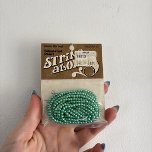 Op de afbeelding: Een pakket vintage imitatieparels in een lichtgroene kleur. De verpakking is voorzien van het opschrift "String Along" en bevat een Sears-prijskaartje. De kralen zijn ongeveer 152 cm lang.