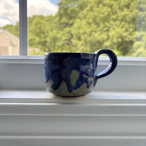 Peut inclure: Une tasse en céramique avec un vernis bleu foncé brillant qui coule sur une base texturée beige clair. La tasse présente une anse arrondie sur le côté droit et un corps cylindrique large avec un bord inférieur légèrement effilé et un rebord lisse.