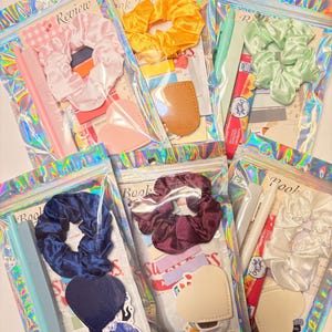 May include: A collection of six iridescent zipper bags, each containing a scrunchie, stickers, and other craft supplies. Scrunchies are in various colors, including pink, yellow, green, blue, burgundy, and white. The bags also contain pens, rulers, and heart-shaped patches.