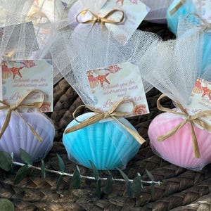 May include: Three shell-shaped bath sponges in lavender, bright blue, and pink, each wrapped in white tulle netting. A tan raffia bow secures a square tag featuring a seashell print and the text Thank you for celebrating with me Love Zara.