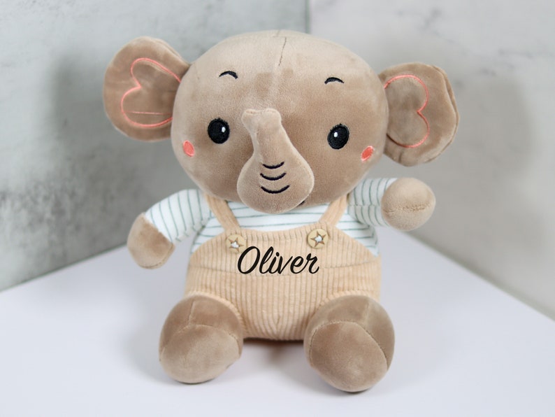 May include: A brown plush elephant toy wearing a striped shirt and overalls. The overalls have a star and the name "Oliver" embroidered on them.