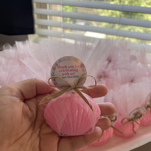 May include: A pink seashell-shaped object wrapped in sheer white tulle fabric, tied with a tan raffia bow. A circular tag is attached, featuring the text "Thank you for celebrating with us! August 23rd 2025" surrounded by small colorful sea-themed illustrations.