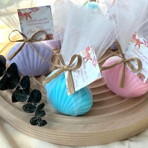 May include: Three seashell-shaped soaps in lavender, bright blue, and pink, each wrapped in white tulle netting and tied with a tan raffia bow. A small square tag with a seashell print and the text Thank you for celebrating with me Love Zara is attached to each soap.