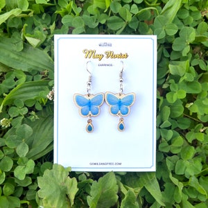 Op de afbeelding: Blauwe vlinder oorbellen met een houten basis en zilveren haakjes. De oorbellen hebben een kleine blauwe traanvormige hanger die aan de onderkant hangt. De oorbellen worden gepresenteerd op een witte kaart met de tekst "Wild & Free May Florist - Earrings - go wildandfree.com"