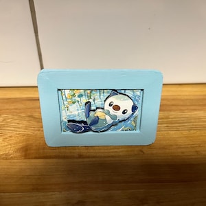 Könnte beinhalten: Ein kleiner, rechteckiger, hellblauer Holzrahmen mit einem geschichteten Papierkunstwerk. Das Bild zeigt eine weiße und blaue Cartoon-Otterfigur, die auf dem Rücken im Wasser treibt und eine gelbe Muschel vor der Brust hält.