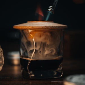 May include: A glass jar with a wooden lid, filled with a dark liquid and white smoke rising from it. A metal tool with a flame is being used to heat the liquid.