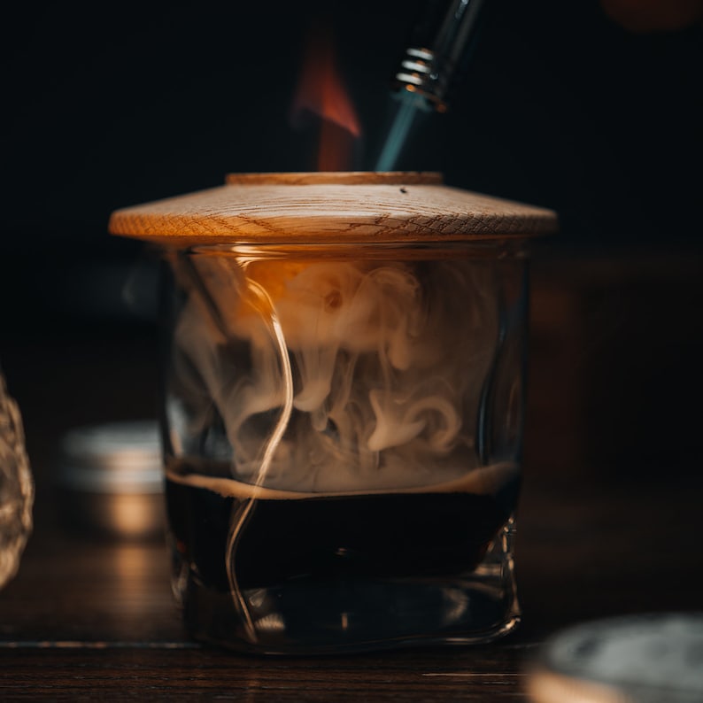 May include: A glass jar with a wooden lid, filled with a dark liquid and white smoke rising from it. A metal tool with a flame is being used to heat the liquid.