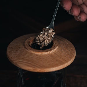 May include: A wooden lid with a small hole in the center is being used to cover a glass jar filled with a dark liquid. A spoon is holding wood chips over the hole in the lid.