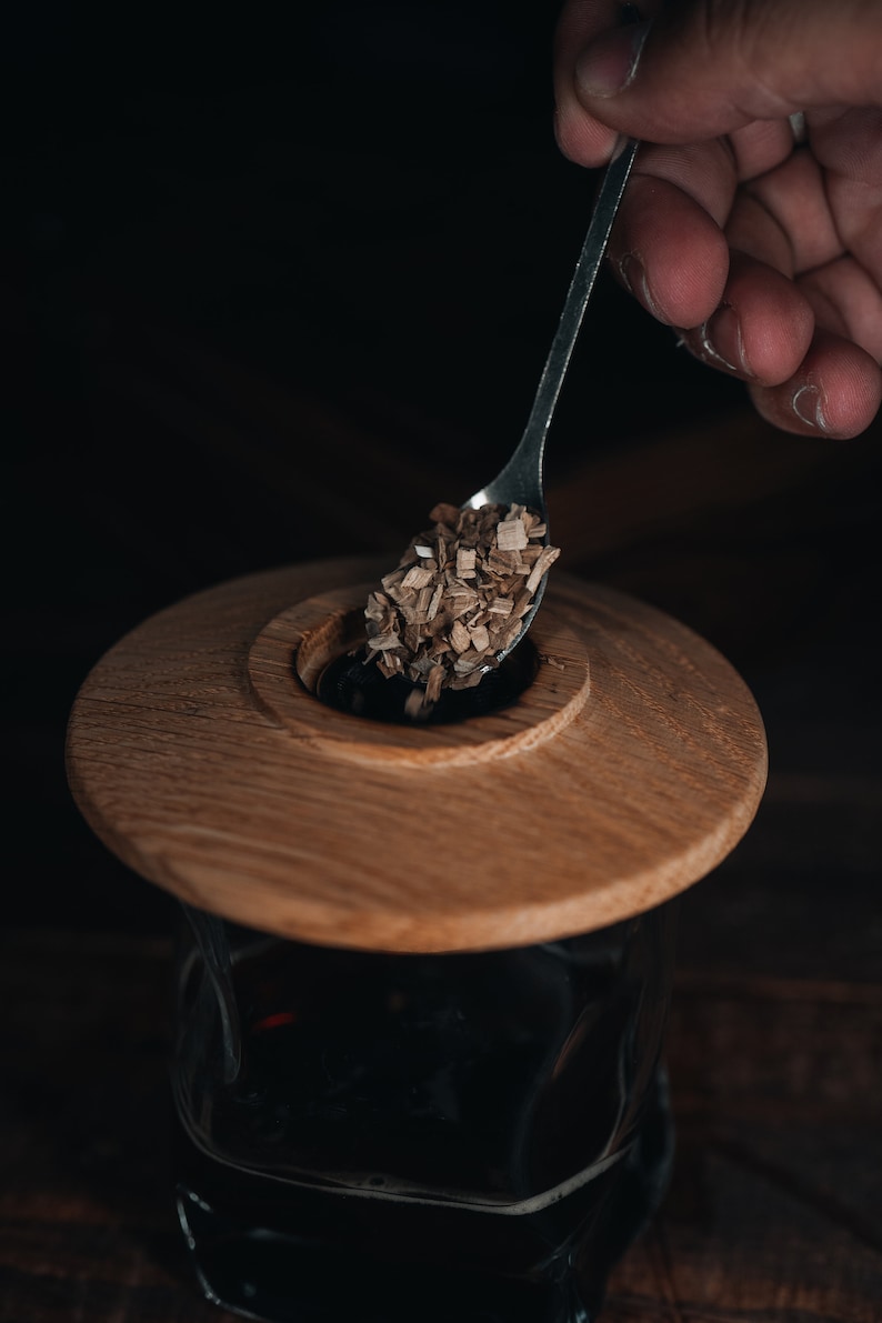 May include: A wooden lid with a small hole in the center is being used to cover a glass jar filled with a dark liquid. A spoon is holding wood chips over the hole in the lid.