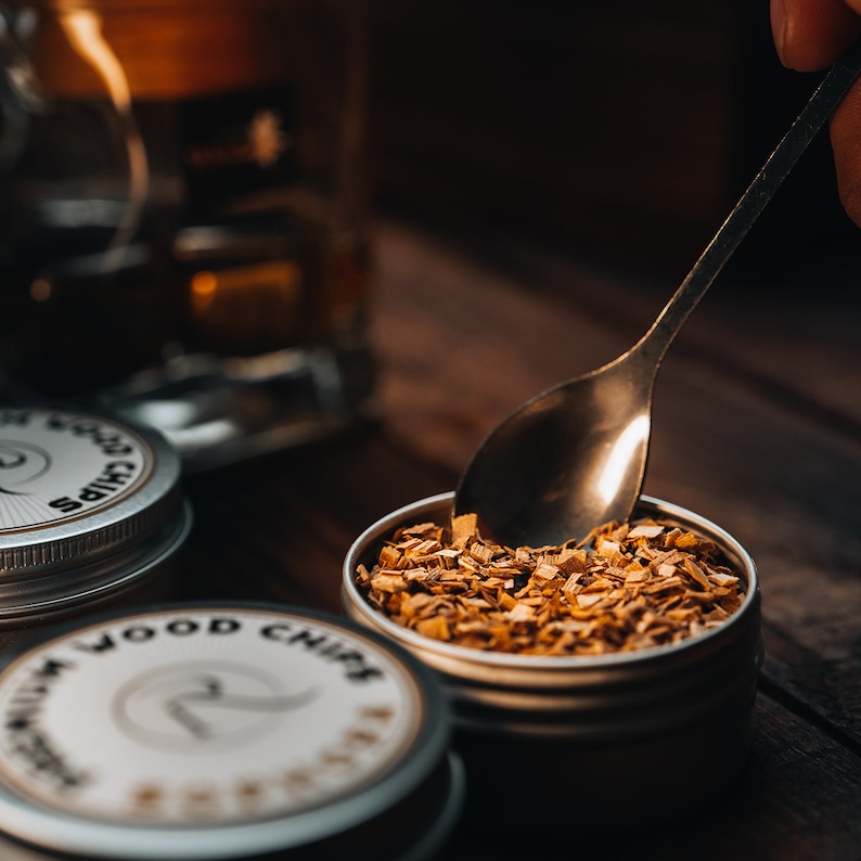 May include: A close-up of a silver tin filled with wood chips. The tin has a white label with the text "Premium Wood Chips" printed on it. A silver spoon is scooping out the wood chips.